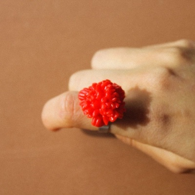 Anel vermelho em forma de flor numa mão com fundo castanho