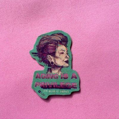 Adesivo com ilustração de mulher idosa e texto em fundo verde sobre superfície rosa.