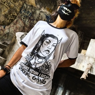 Pessoa com t-shirt branca estampada, máscara preta com dentes, óculos escuros em ruína com grafitis