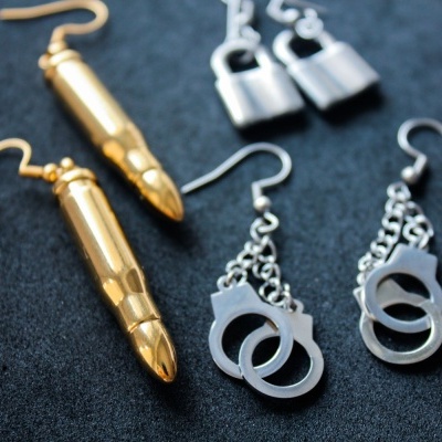 Brincos dourados em forma de bala e prateados com algemas em fundo preto.