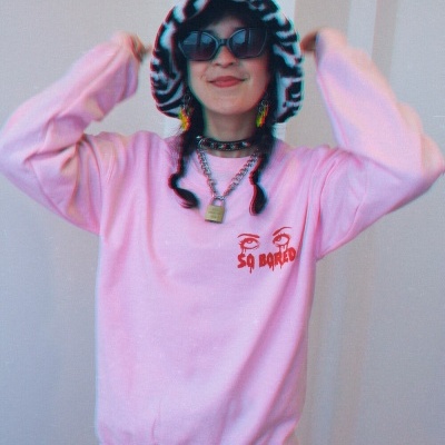 Pessoa com camisola rosa e chapéu estampado segurando chapéu, com calções quadriculados e óculos escuros