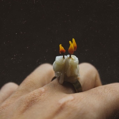 Anel com velas pequenas derretidas e chamas laranja num dedo, fundo preto.