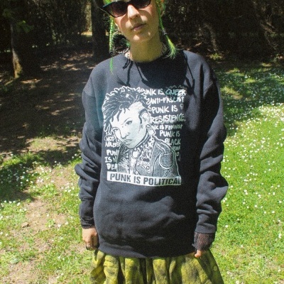 Pessoa com sweat preta com texto punk e saia xadrez verde em ambiente exterior
