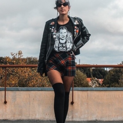 Pessoa com casaco de cabedal preto, t-shirt preta com texto Eat the RICH, saia xadrez, meias pretas e óculos escuros numa área exterior