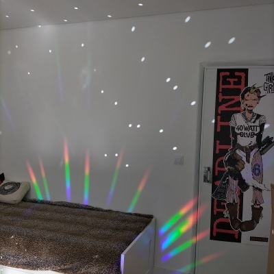 Quarto com cama branca, manta castanha, reflexos de luz coloridos na parede e porta com poster de banda desenhada