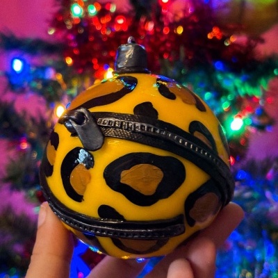 Bola de Natal amarela com padrão de pele de leopardo e fecho de zíper em frente a árvore de Natal com luzes coloridas