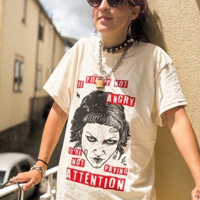 T-shirt bege com impressão de rosto masculino e texto em vermelho e branco usada por pessoa com óculos escuros e cabelo roxo