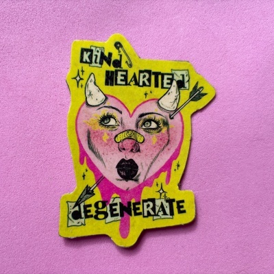 Adesivo de coração rosa com rosto, chifres e flechas em fundo amarelo e texto preto, sobre fundo rosa.