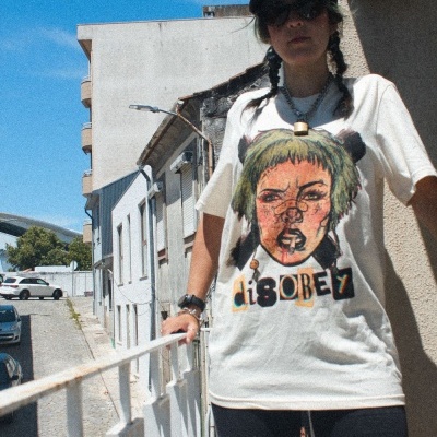 Pessoa com t-shirt branca estampada e calções pretos junto a uma grade branca numa rua urbana