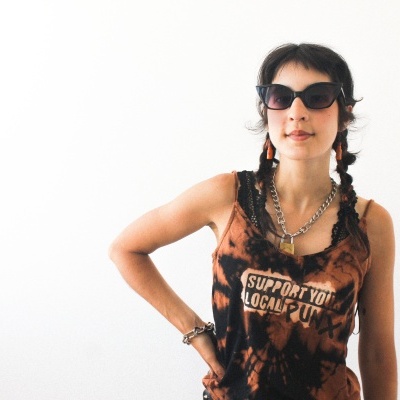 Pessoa com regata tie-dye castanho e preto com texto, óculos de sol pretos e acessórios prateados e vermelhos, fundo branco