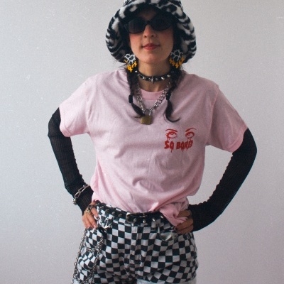 Pessoa com chapéu e roupa estilosa com padrão quadriculado e texto SO BORED na t-shirt