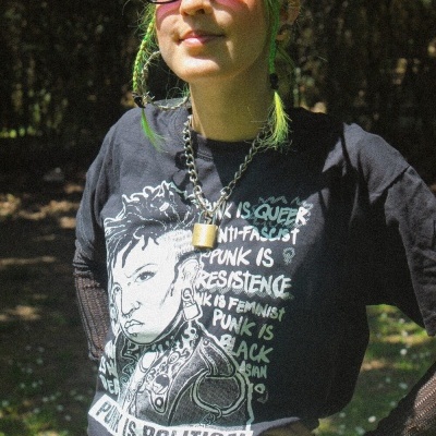 Pessoa com t-shirt preta com texto e ilustração, cabelo verde e óculos escuros