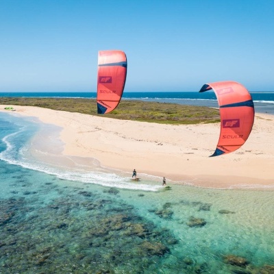 Dois kitesurf vermelhos a voar sobre mar azul claro junto a uma ilha arenosa com vegetação