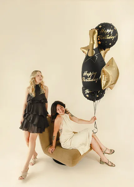 Conjunto de balões Garrafa Happy Birthday Duas mulheres com vestidos elegantes e balões de Happy Birthday num fundo neutro.
