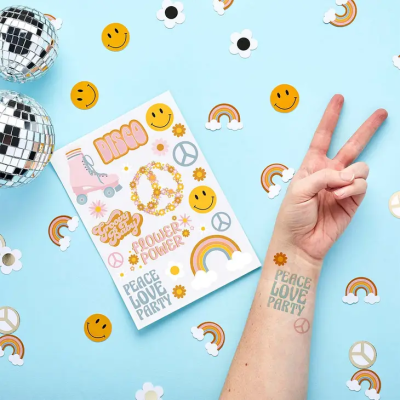 Braço com tatuagem temporária e conjunto de tatuagens com temas disco, flores e arco-íris sobre fundo azul claro decorado.