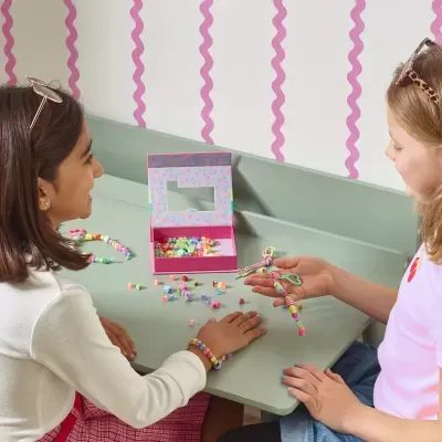 Duas raparigas a fazer pulseiras com miçangas coloridas sentadas numa mesa verde clara com caixa rosa e parede branca com listas rosa atrás.