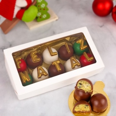 Caixa de bombons decorados com letras douradas formando FELIZ NATAL com bombons cortados ao lado e decoração natalícia ao fundo