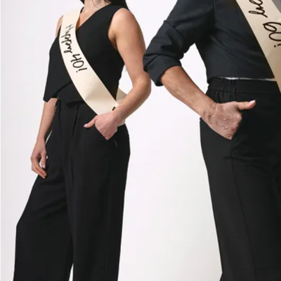 Duas pessoas com roupas pretas usando faixas creme com texto comemorativo, fundo branco.