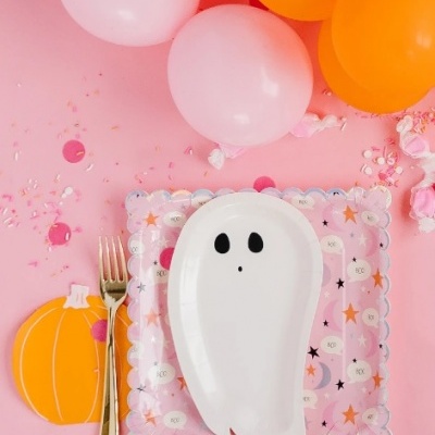 Prato em forma de fantasma branco sobre guardanapo com estrelas e luas, garfo dourado, balões laranja e rosa e abóbora em fundo rosa