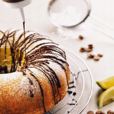 Bolo redondo com cobertura de açúcar em pó e calda de chocolate a ser vertida, limão e grãos de café ao fundo