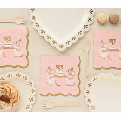 Conjunto de festa com guardanapos cor-de-rosa com ratinhos, pratos brancos com bordas douradas e talheres de madeira.