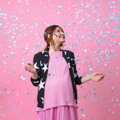 Mulher com casaco preto de estrelas brancas e vestido rosa plissado rodeada por confetes azuis