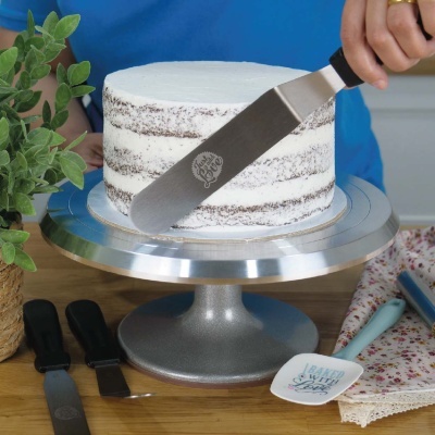 Bolo branco com camadas, base metálica e utensílios de cozinha para decorar