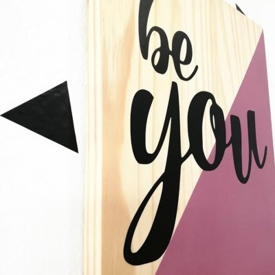 Placa decorativa em madeira com texto 'be you' e seta preta na parede