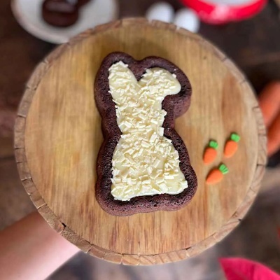 Bolo em forma de coelho de chocolate com cobertura branca sobre tábua de madeira redonda e mini cenouras decorativas
