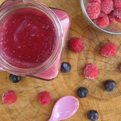 Frasco com puré vermelho, colher rosa, framboesas e mirtilos sobre madeira