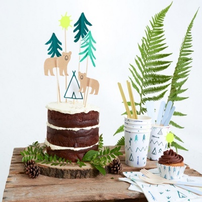 Bolo de chocolate decorado com toppers de papel de ursos e árvores, rodeado por elementos naturais e artigos de festa com padrão de natureza