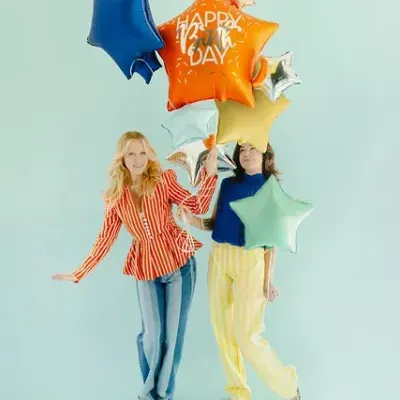 Duas mulheres com balões metalizados em forma de estrelas coloridas, fundo azul claro
