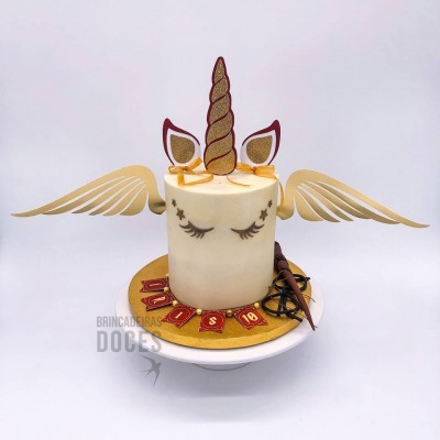 Bolo branco decorado com chifres e orelhas de unicórnio dourados com brilhos e asas douradas laterais.