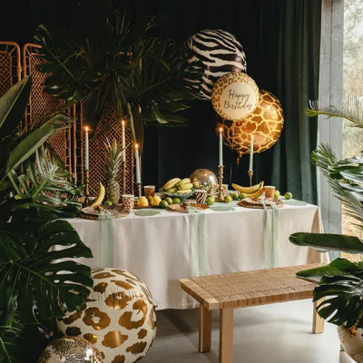 Mesa de festa decorada com toalha branca, velas acesas e balões animal print Happy Birthday em ambiente tropical