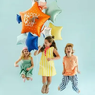Três crianças a saltar com balões em forma de estrela coloridos e um balão laranja com texto HAPPY BIRTHDAY em fundo azul claro.