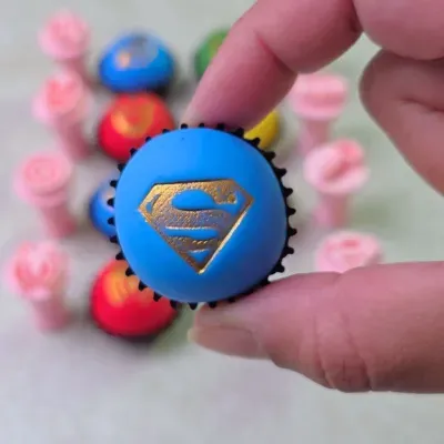 Cupcake azul com símbolo dourado do Superman segurado por mão com outras peças coloridas ao fundo
