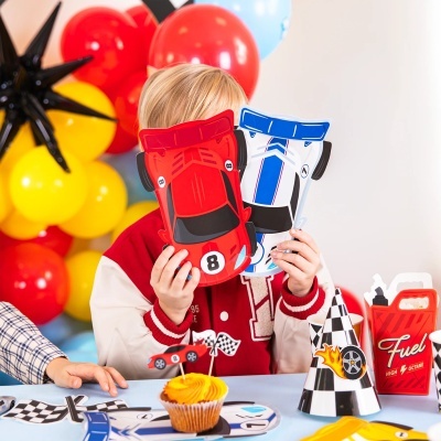 Criança segura cartões de carros de corrida em festa temática com balões, cupcake e decoração de corrida.
