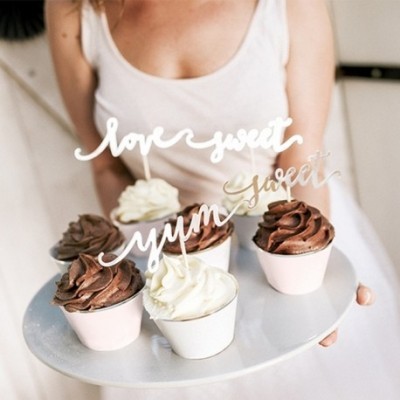 Bandeja com cinco cupcakes decorados com toppers de texto festivo, segurada por pessoa de camisola branca.