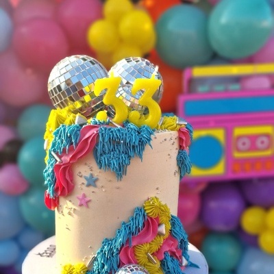 Bolo decorado com velas 33 e bolas espelhadas num suporte roxo com balões coloridos ao fundo