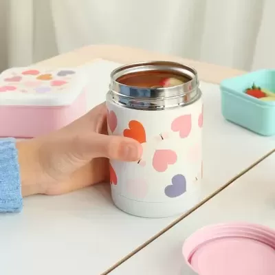 Frasco térmico branco com corações coloridos e tampa rosa, segurado por mão com suéter azul, sobre mesa branca com recipientes coloridos ao fundo.