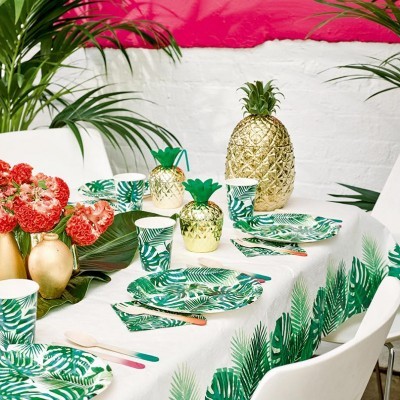 Mesa decorada com tema tropical, pratos e copos com padrão verde, jarros abacaxi dourados, talheres coloridos e flores vermelhas.