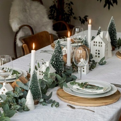 Mesa posta decorada com velas, árvores de Natal artificiais, casas decorativas e pratos com bases de madeira