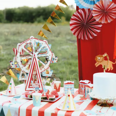 Mesa de festa ao ar livre com decoração colorida incluindo bolo, chapéus de festa, roda-gigante de papel e guirlanda dourada.