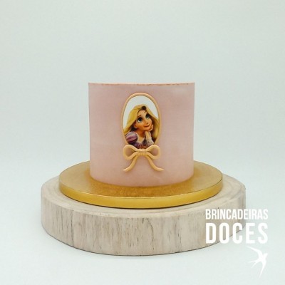 Bolo pequeno rosa com decoração da Rapunzel e laço, em prato dourado sobre base de madeira