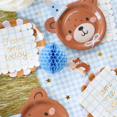Pratos de papel com urso castanho e placas decorativas com texto 'I am one today' sobre toalha azul xadrez
