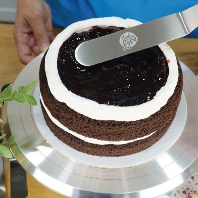 Bolo de chocolate com cobertura branca e geleia escura sendo espalhada com espátula metálica. 