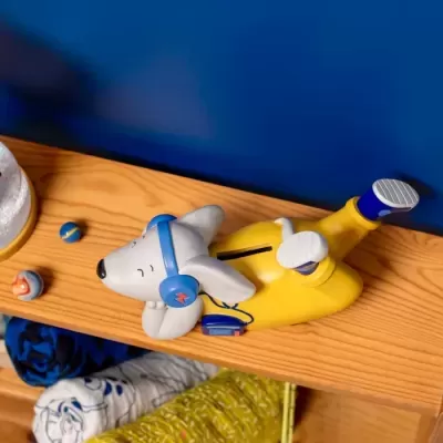 Figura decorativa de cão deitado com auscultadores e fato amarelo numa prateleira de madeira com bolas coloridas e têxteis.