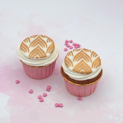 Dois cupcakes em forminhas rosa com decoração branca e bolacha decorada em cima, com confeites rosa ao redor