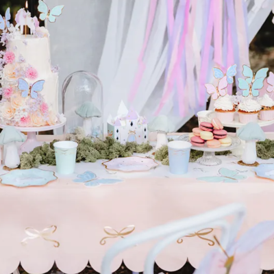 Mesa de festa decorada com bolo, cupcakes, macarons e toalha rosa, cenário em tons pastel.