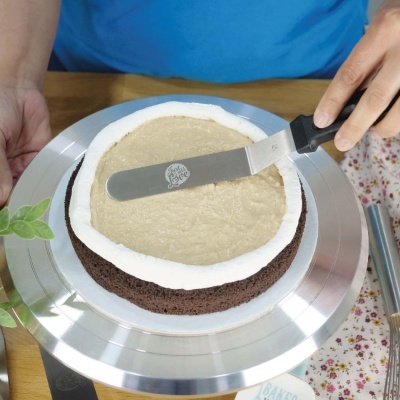 Bolo de chocolate com cobertura branca e creme sendo decorado com espátula.